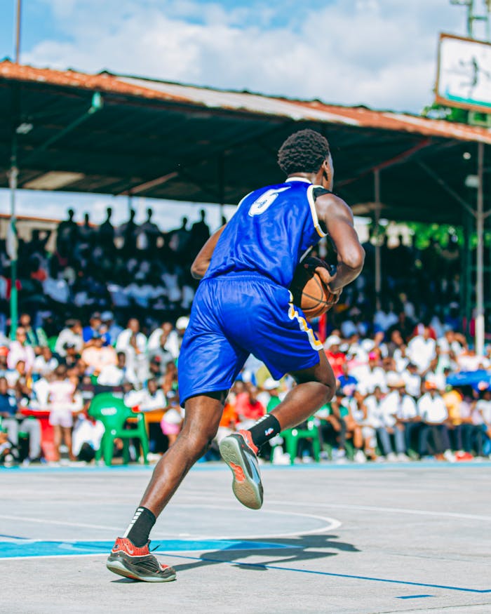 Basketball player dribbles on outdoor court with cheering audience under sunny sky.
