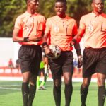 Three soccer referees in orange uniforms walking on a sunny field.