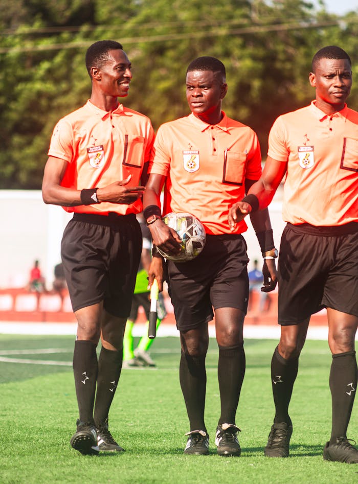 Three soccer referees in orange uniforms walking on a sunny field.